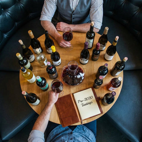 Two people tasting wine around a round wooden table with wine bottles and glasses.